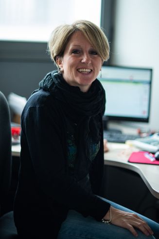 Una donna con i capelli biondi corti sorride, indossa un maglione nero e una sciarpa ed è seduta a una scrivania vicino a un computer.