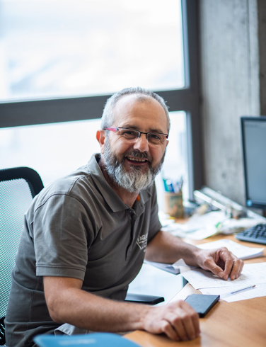 Un uomo con la barba grigia e gli occhiali sorride alla scrivania, tenendo in mano dei documenti, vicino a una finestra.