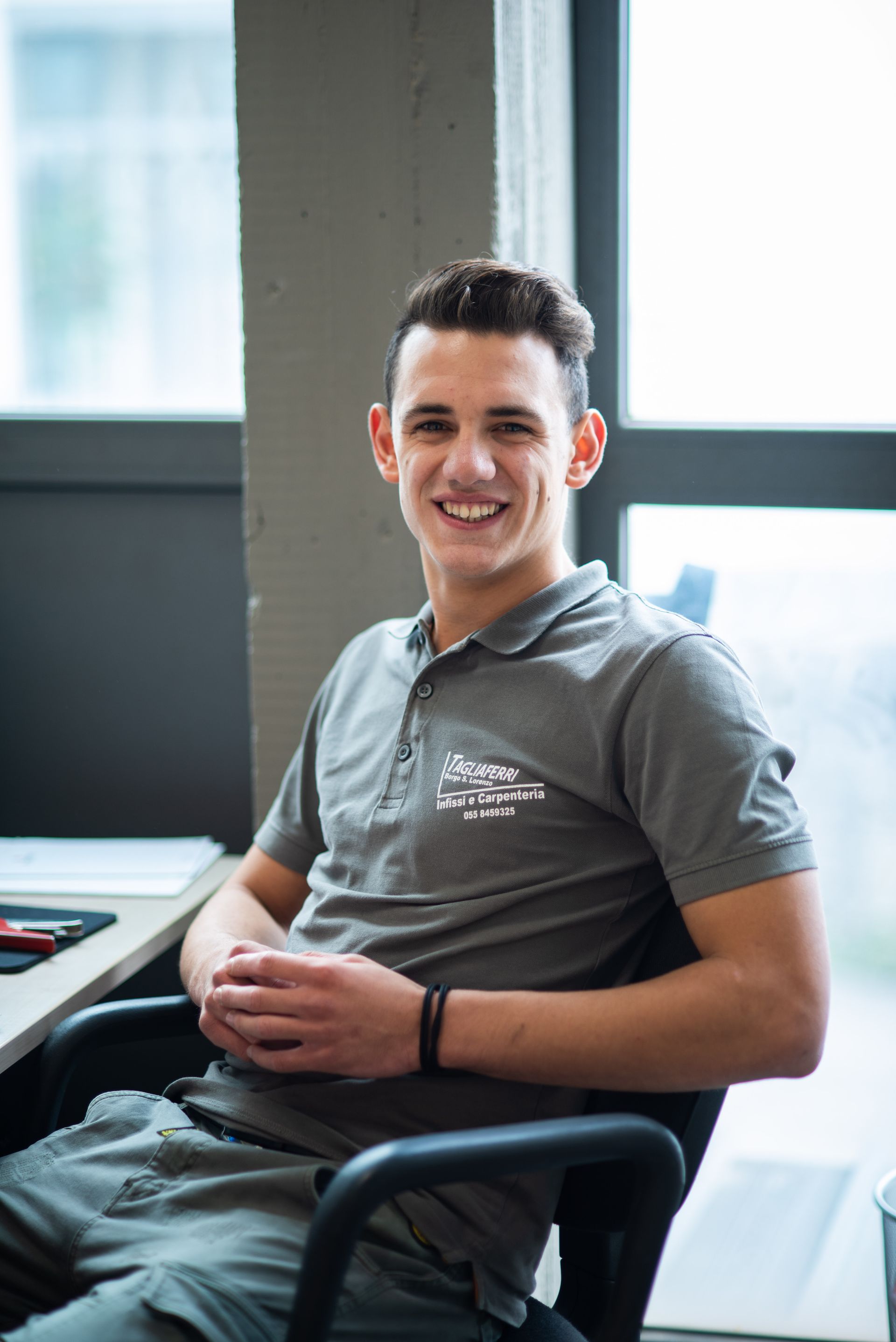 Un uomo con una polo grigia sorride, seduto a una scrivania vicino a una finestra, con le braccia incrociate.
