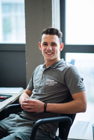 Un uomo sorridente indossa una polo grigia ed è seduto su una sedia da ufficio vicino a una finestra.