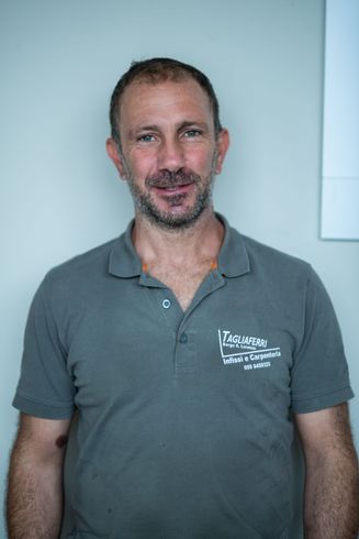 Uomo sorridente con indosso una polo grigia, in piedi contro un muro grigio chiaro.