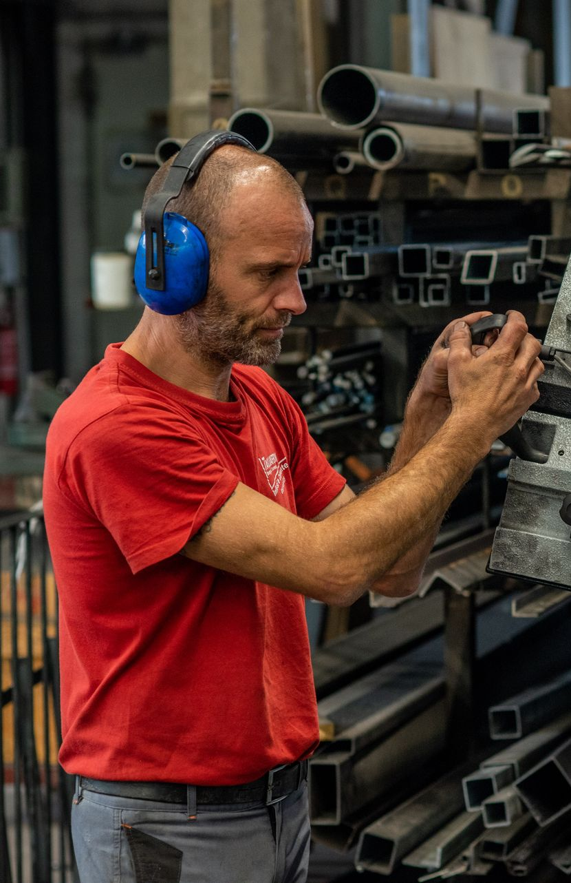 Un uomo con una camicia rossa e le cuffie antirumore esamina dei pezzi di metallo in un'officina.