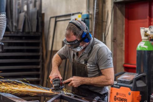 Un uomo indossa un equipaggiamento di sicurezza e macina il metallo in un'officina, mentre scoccano scintille.