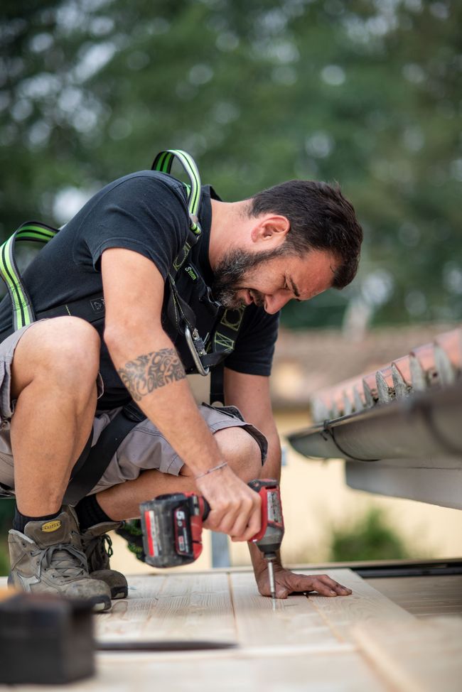 Un operaio edile si accovaccia sul tetto e sta perforando. Indossa un'imbracatura e degli stivali da lavoro, e ha un tatuaggio.