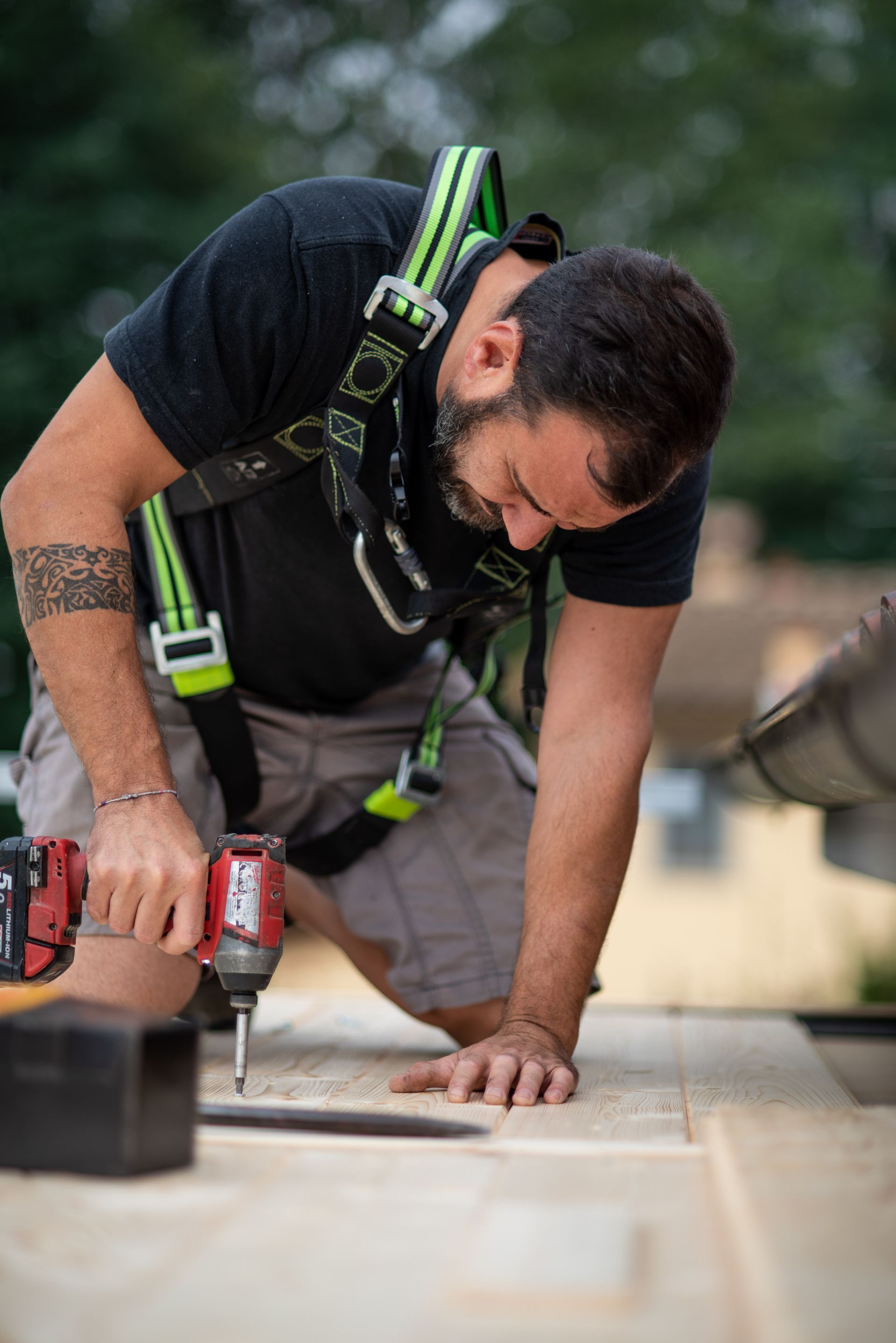 Uomo con barba e imbracatura di sicurezza che usa un trapano su una superficie di legno all'aperto.
