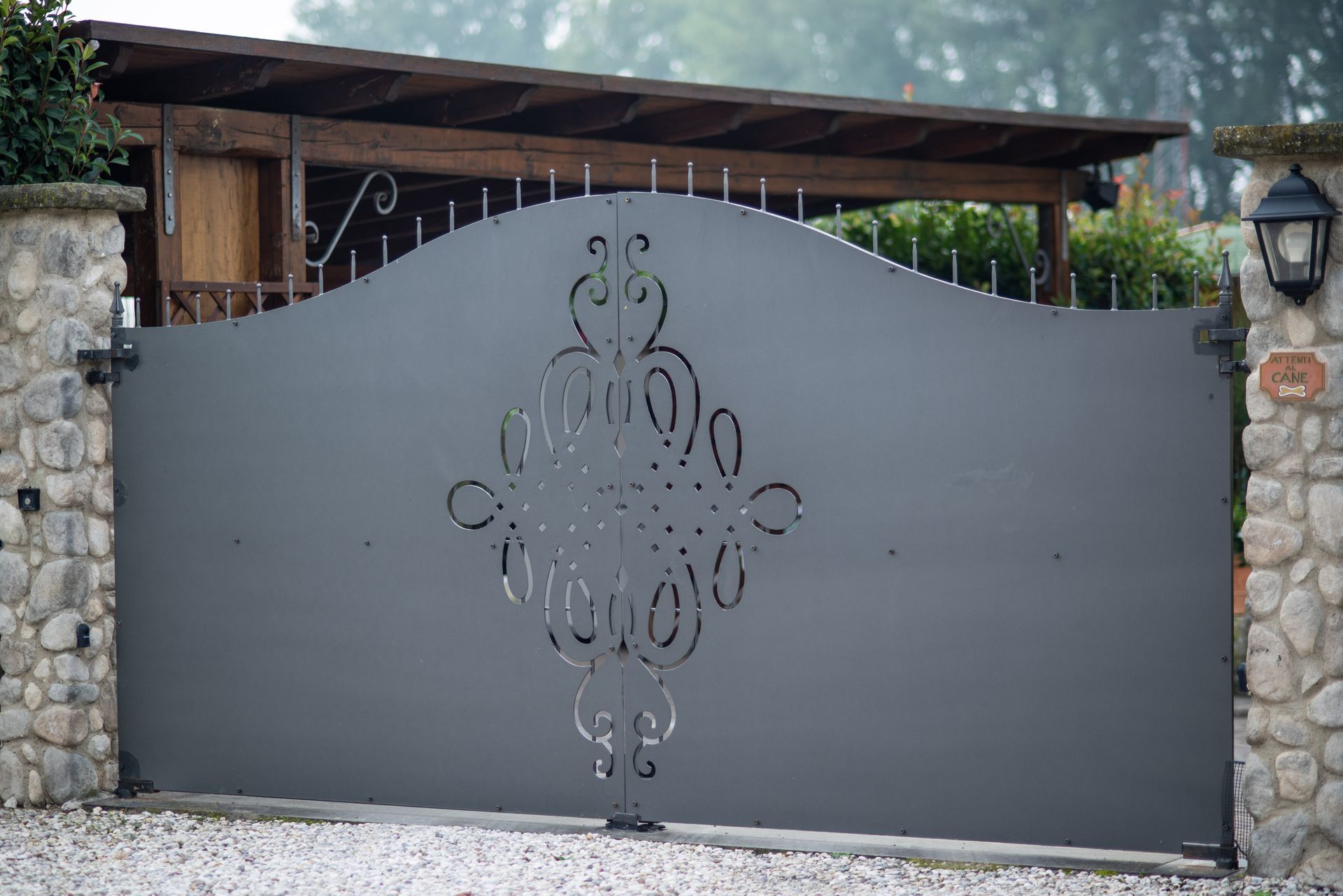 Cancello in metallo grigio con ritagli decorativi, pilastri in pietra e un riparo in legno sullo sfondo.