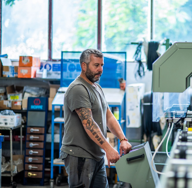 Uomo che lavora in un'officina, azionando macchinari. Ha tatuaggi ed è concentrato.