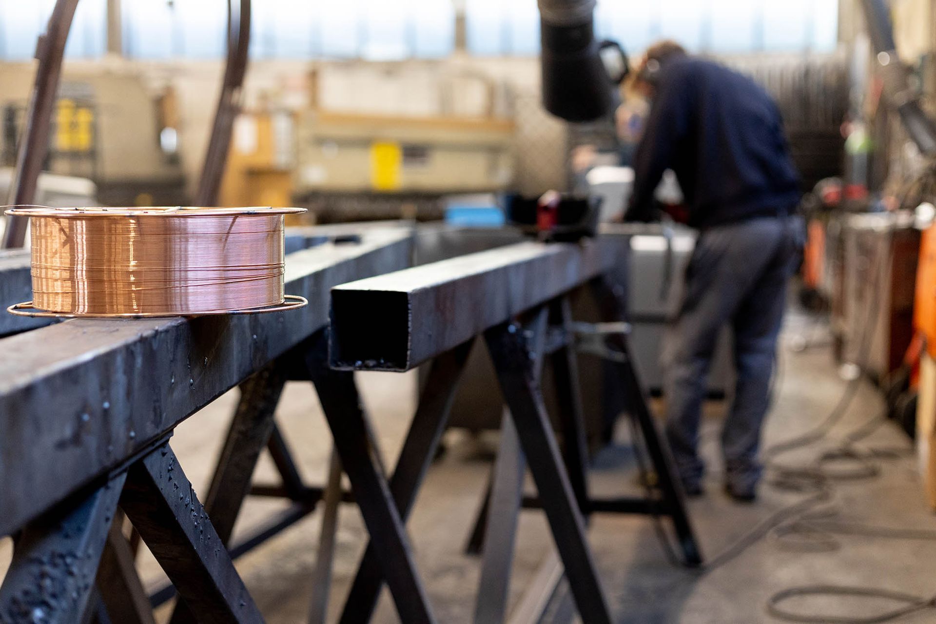 Bobina di filo di rame su trave metallica, operaio sullo sfondo che taglia il metallo, officina.