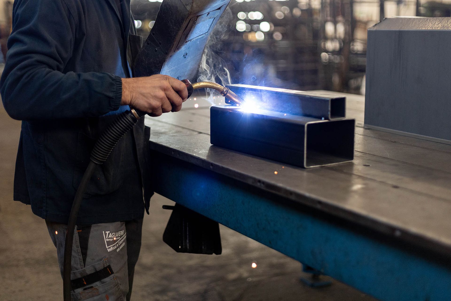 Saldatore che utilizza la torcia su travi metalliche su un banco da lavoro, producendo scintille luminose in un ambiente industriale.
