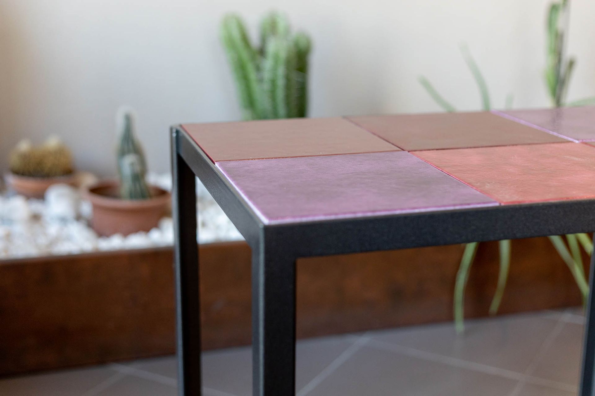 Tavolino con struttura in metallo nero e piano in piastrelle quadrate nei toni del rosso e del viola. Cactus sullo sfondo.