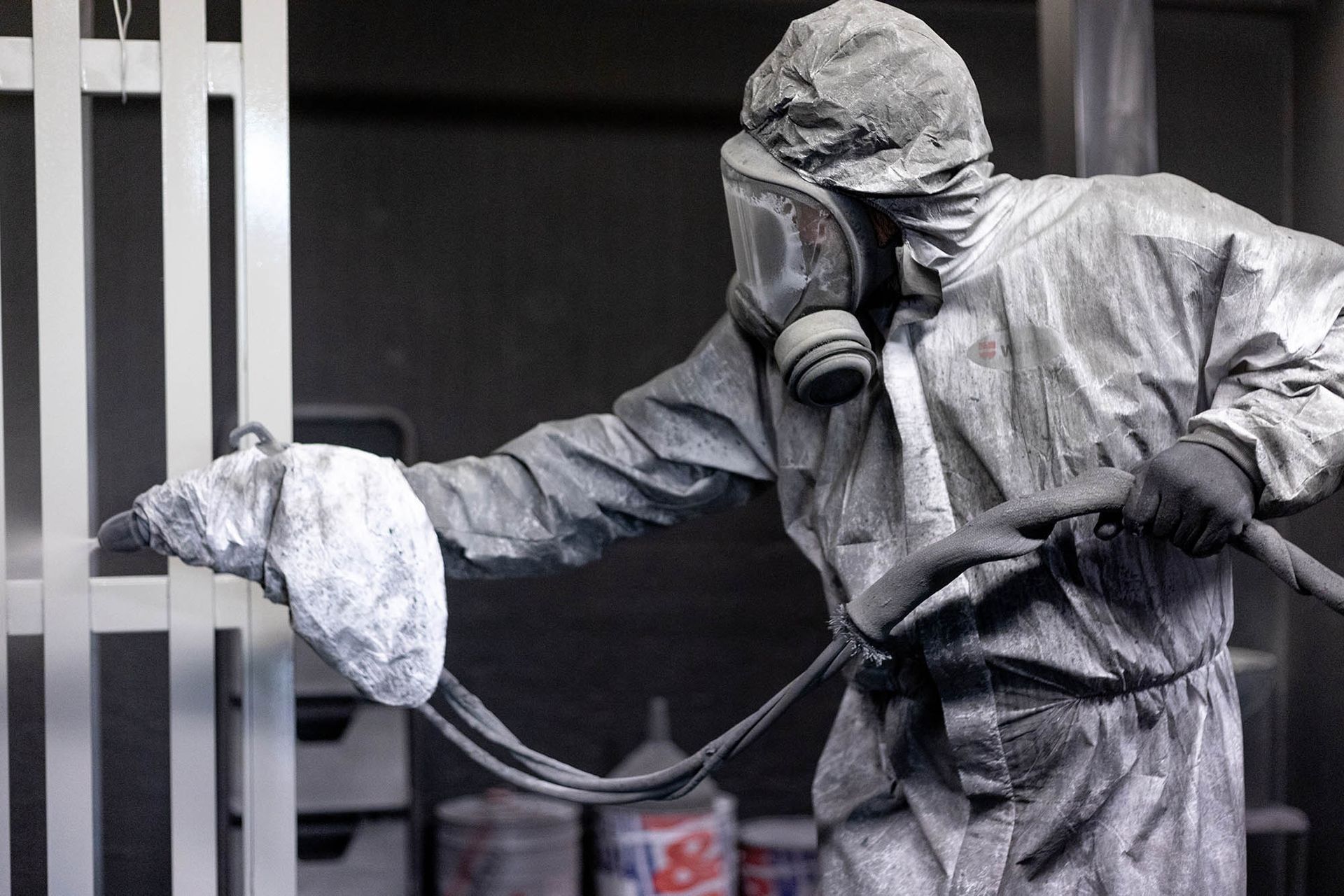 Persona con tuta protettiva e maschera antigas che vernicia a spruzzo una struttura metallica in un ambiente interno.