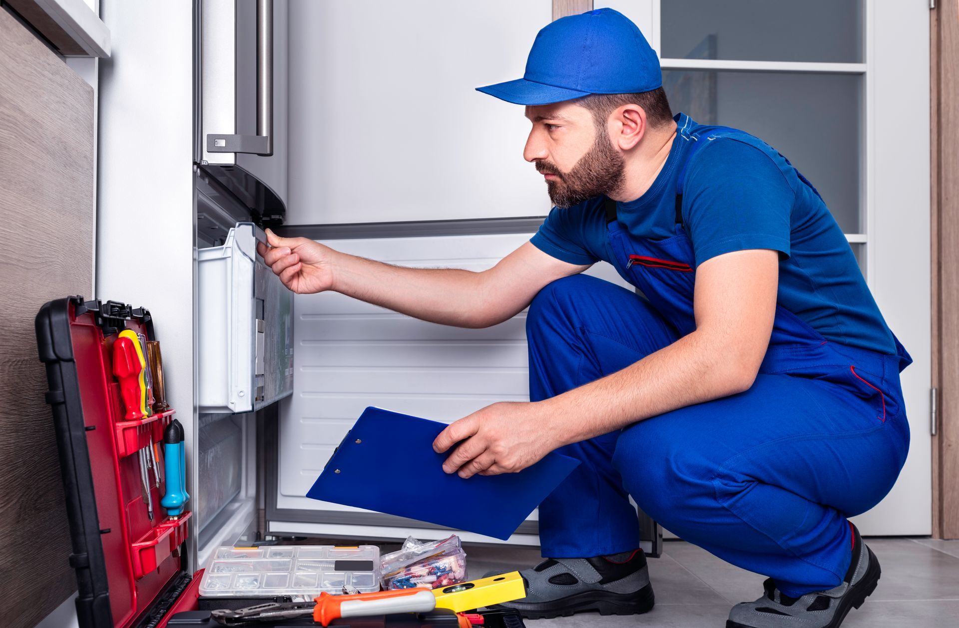 A man is kneeling down in front of a refrigerator while holding a clipboard.