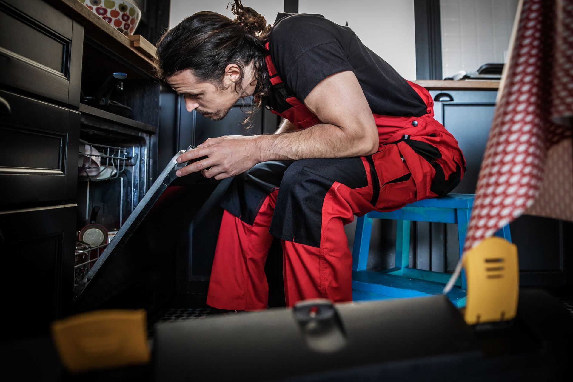 A man in red overalls is working on a dishwasher in a kitchen.