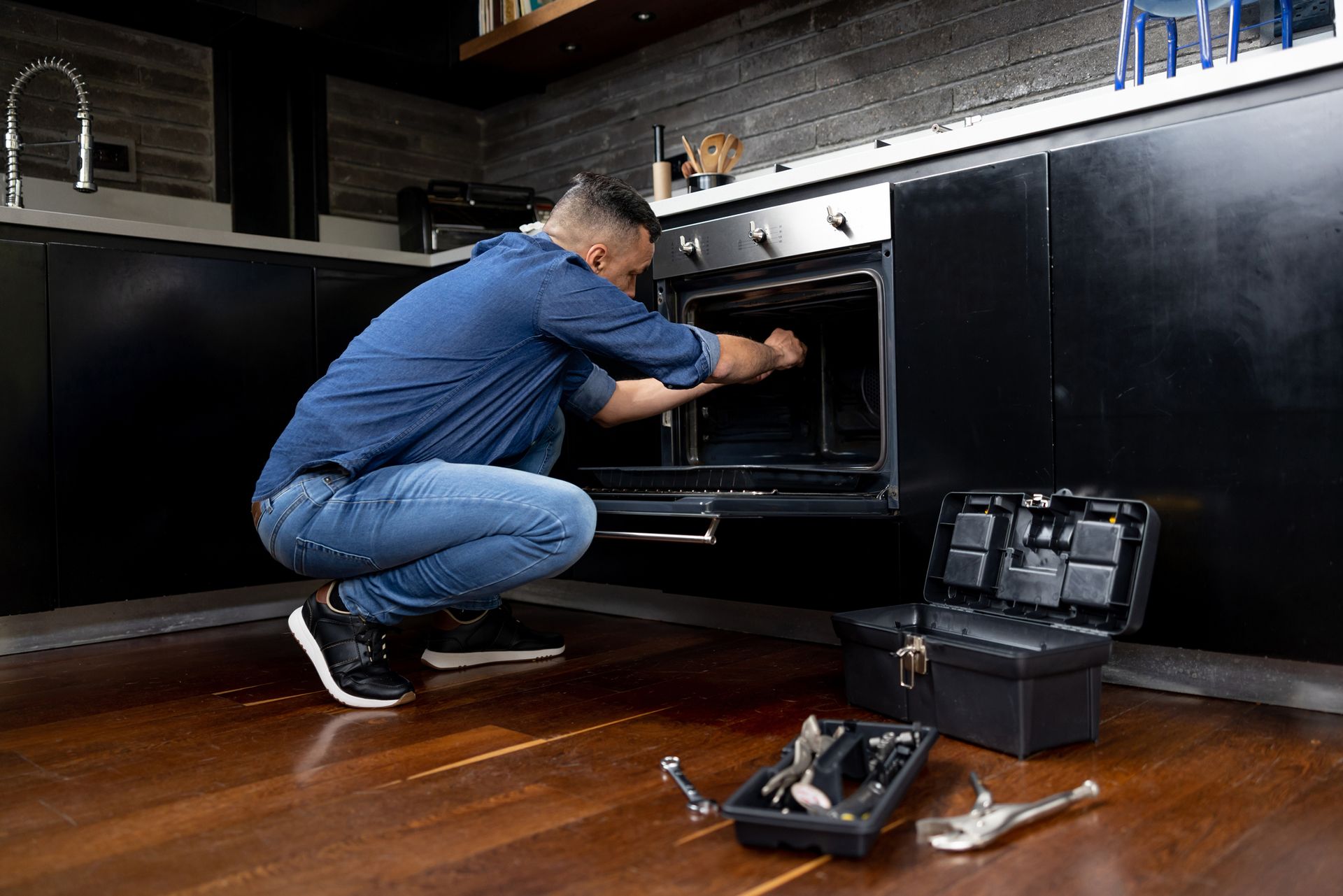 A man is kneeling down in a kitchen fixing an oven.