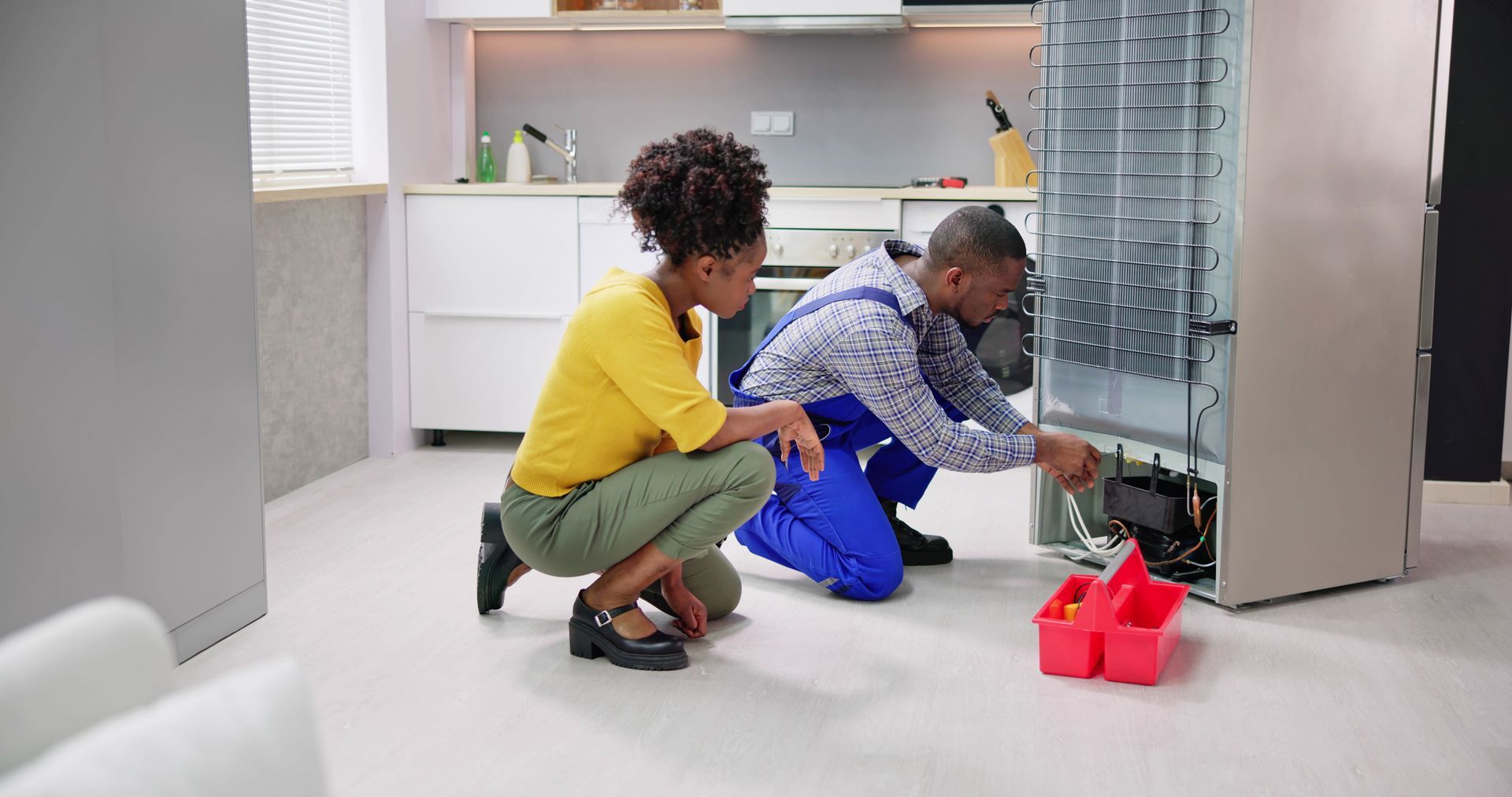 A man and a woman are working on a refrigerator in a kitchen.