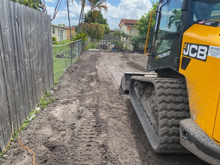 A yellow bulldozer is driving down a dirt road next to a wooden fence.