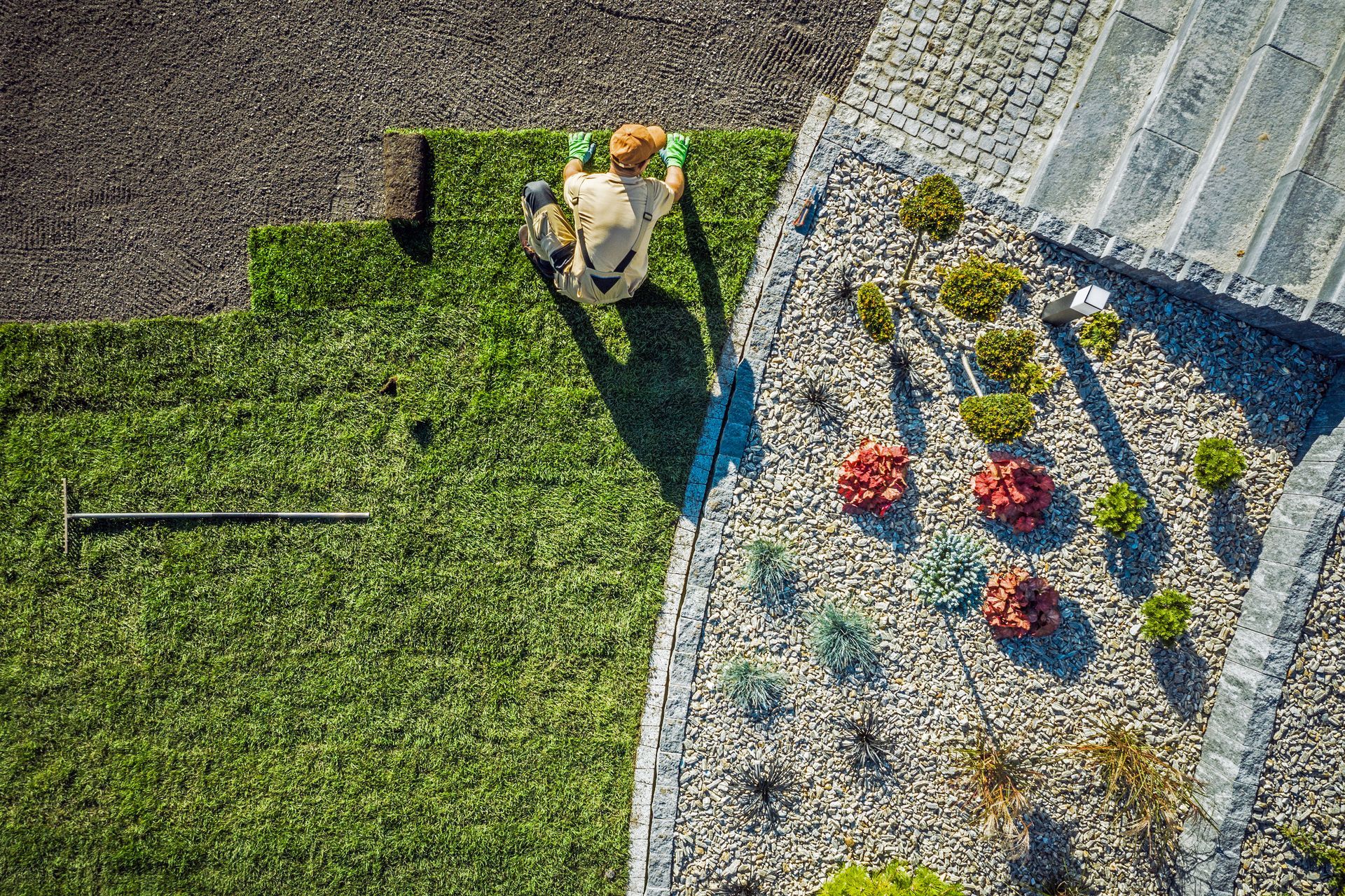 Top view of a landscaper laying fresh sod next to decorative gravel and plants in a garden design.