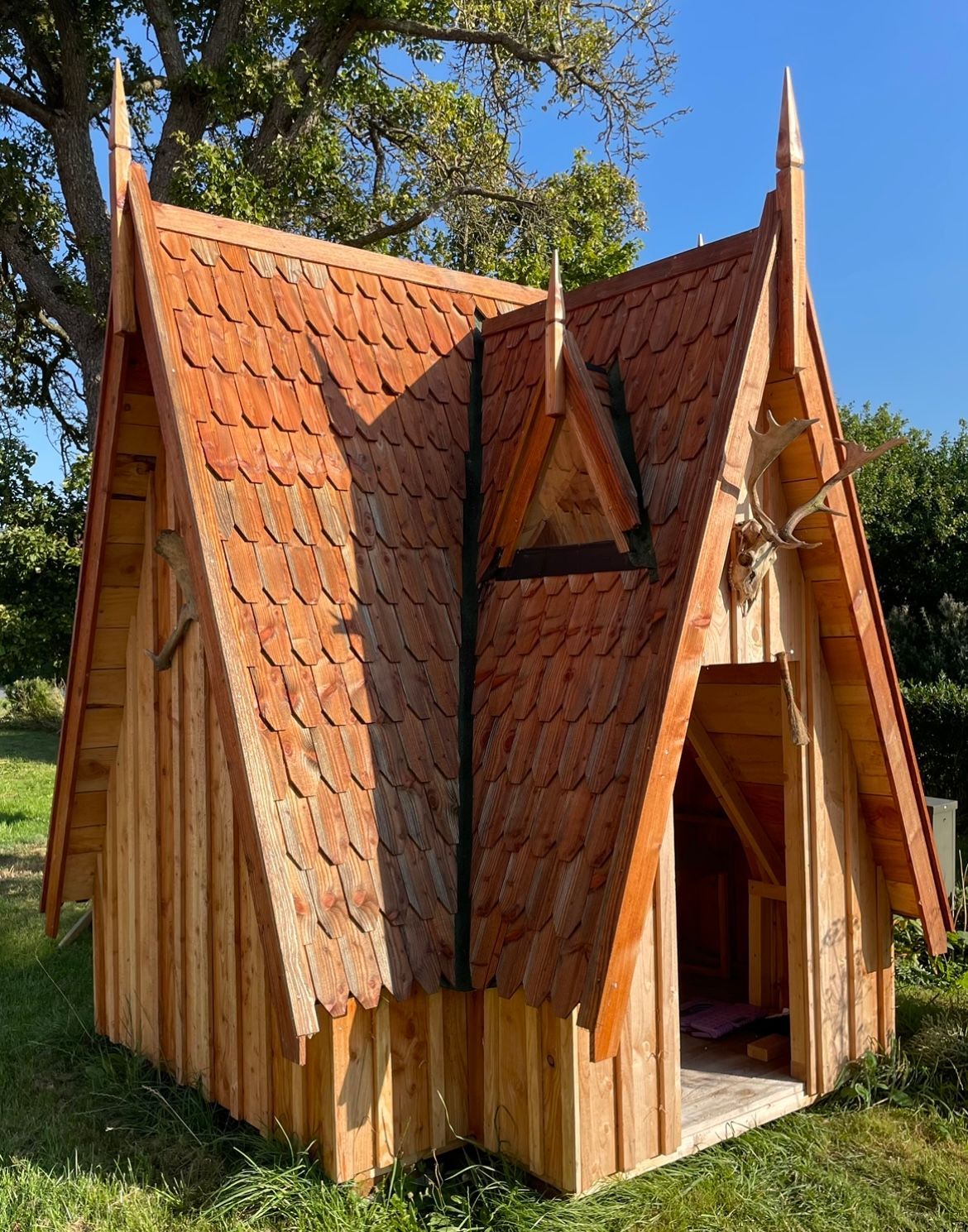 lille træbygning, der ligner et eventyrhus eller en miniatureudgave af et vikingehus. Taget er dækket af håndskårne træspåner i skælleform og har en markant, stejl hældning med flere spidse gavle og dekorative spidser i toppen. Facaden og væggene er lavet af lodrette træplanker, og huset har en åben indgang uden dør. 