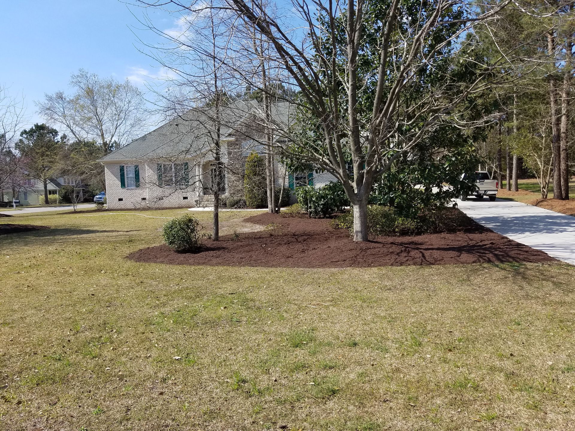 A house with tan brick siding, green shutters, and a gravel driveway. Brown mulch surrounds trees and bushes on the lawn.