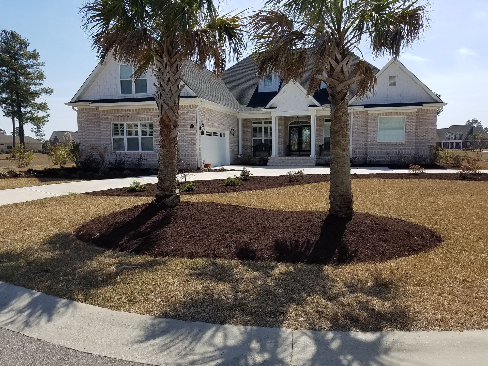 A brick house with palm trees in front, a circular flower bed with mulch, and a driveway.