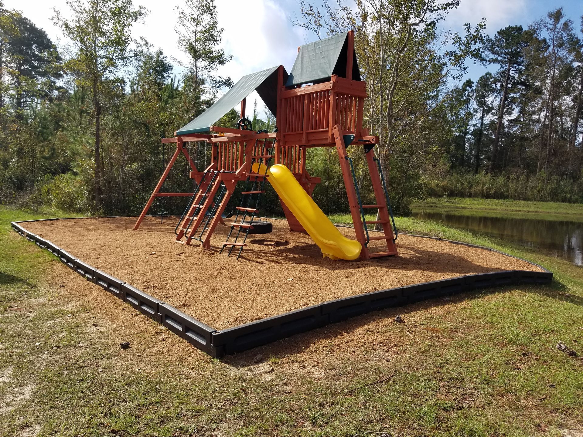 Wooden playground set with slide and climbing ladders on mulch bed near trees and a pond.