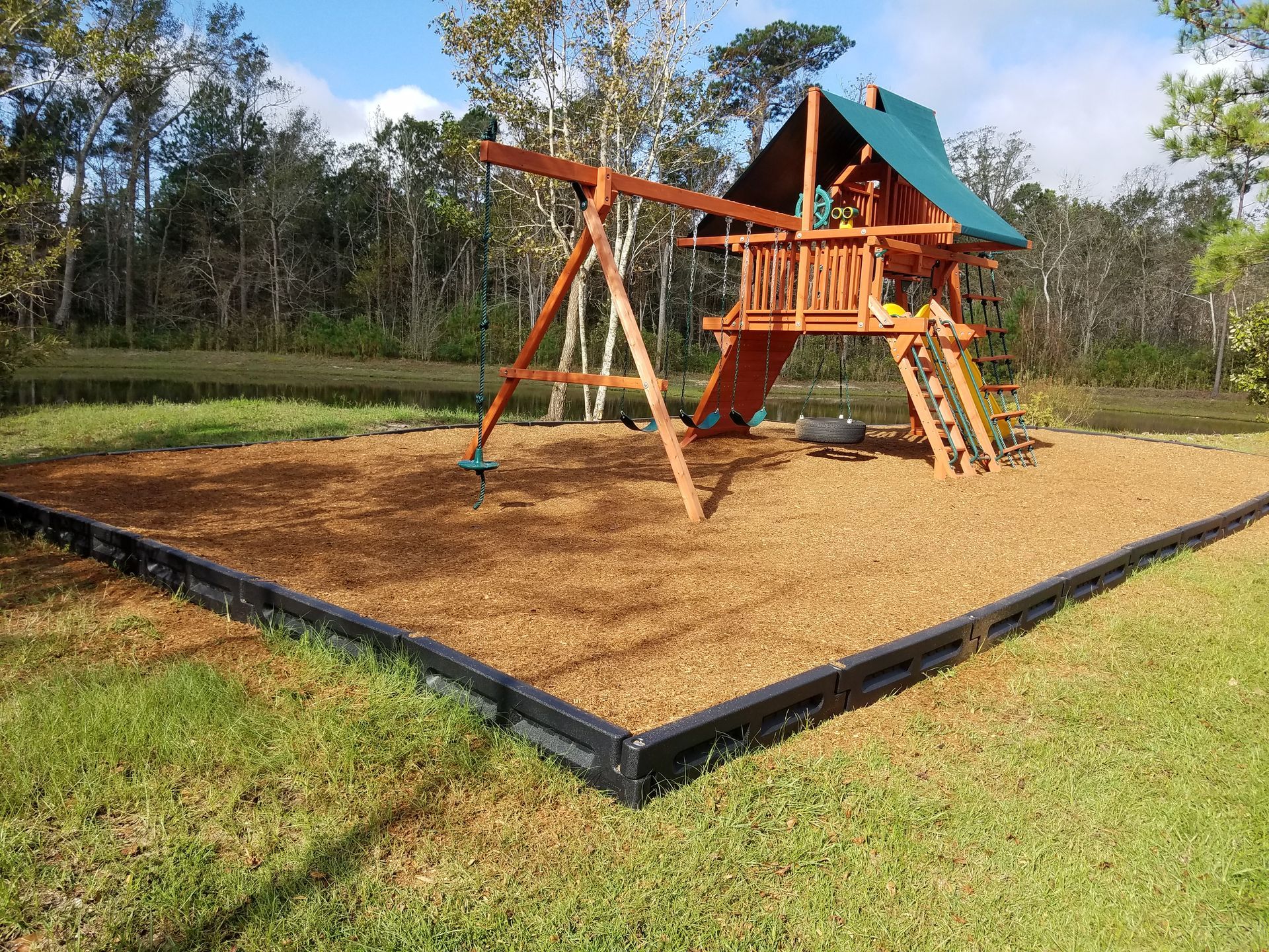 Playground with wooden swing set and slide on woodchip base, surrounded by grass.