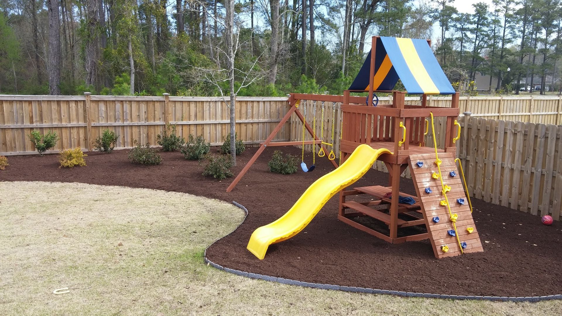 Playground with yellow slide, climbing wall, and swing set, on brown mulch, near a wooden fence.