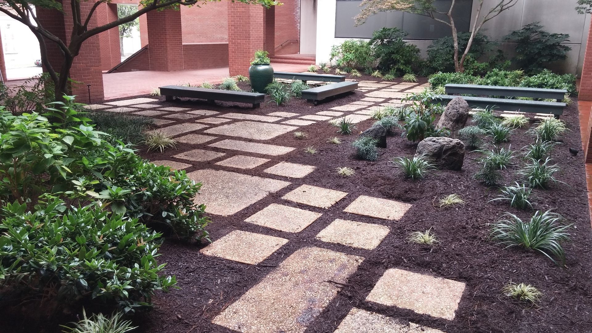 Stone path through a Japanese garden with green plants, dark mulch, and brick building.