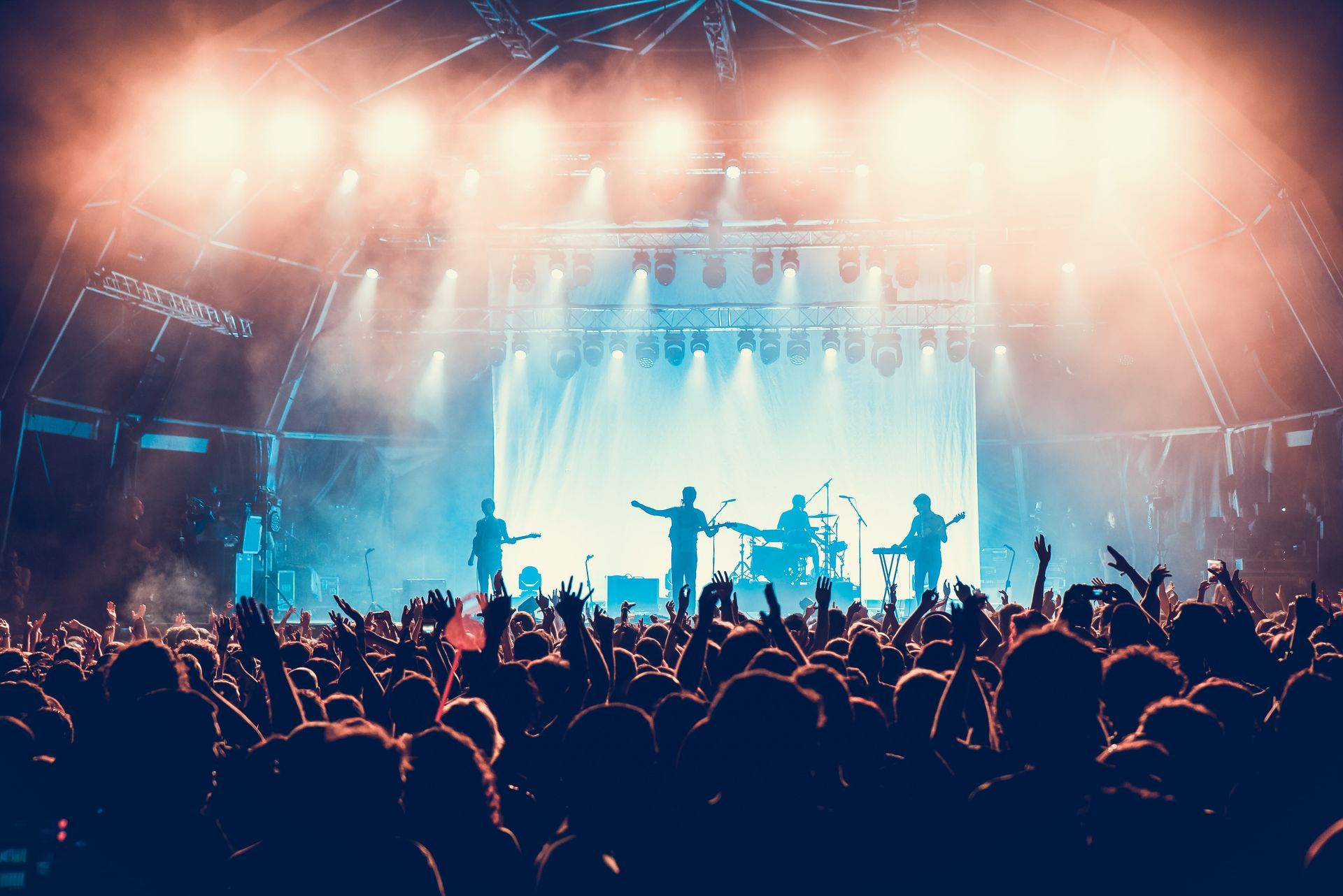 Band performing on stage, backlit by bright lights, in front of a cheering crowd with raised hands.