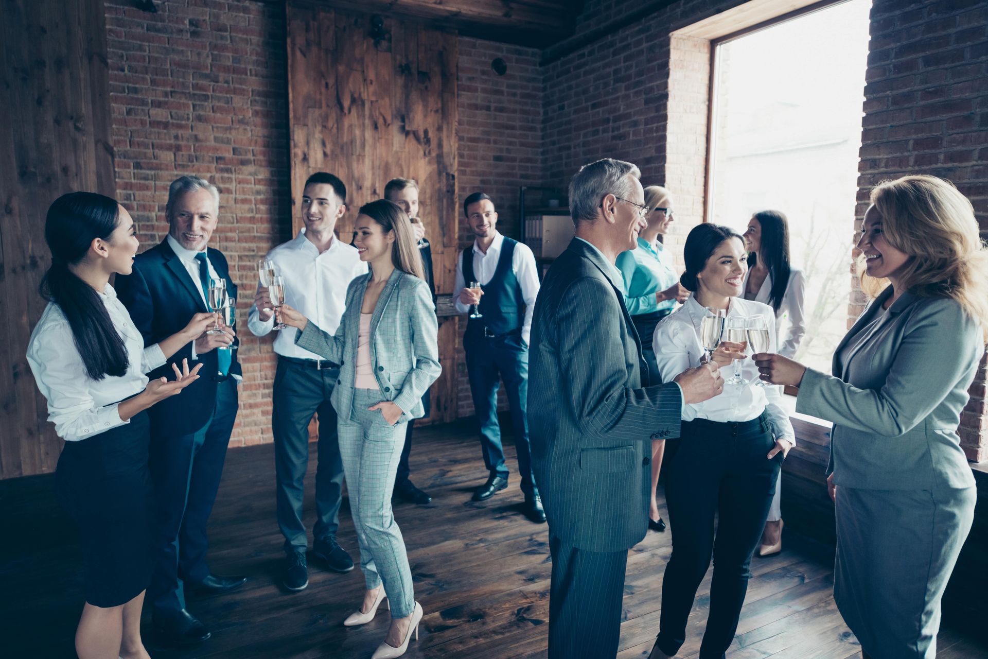 People in business attire at a networking event in a room with brick walls. Toasting and conversing, some with drinks.
