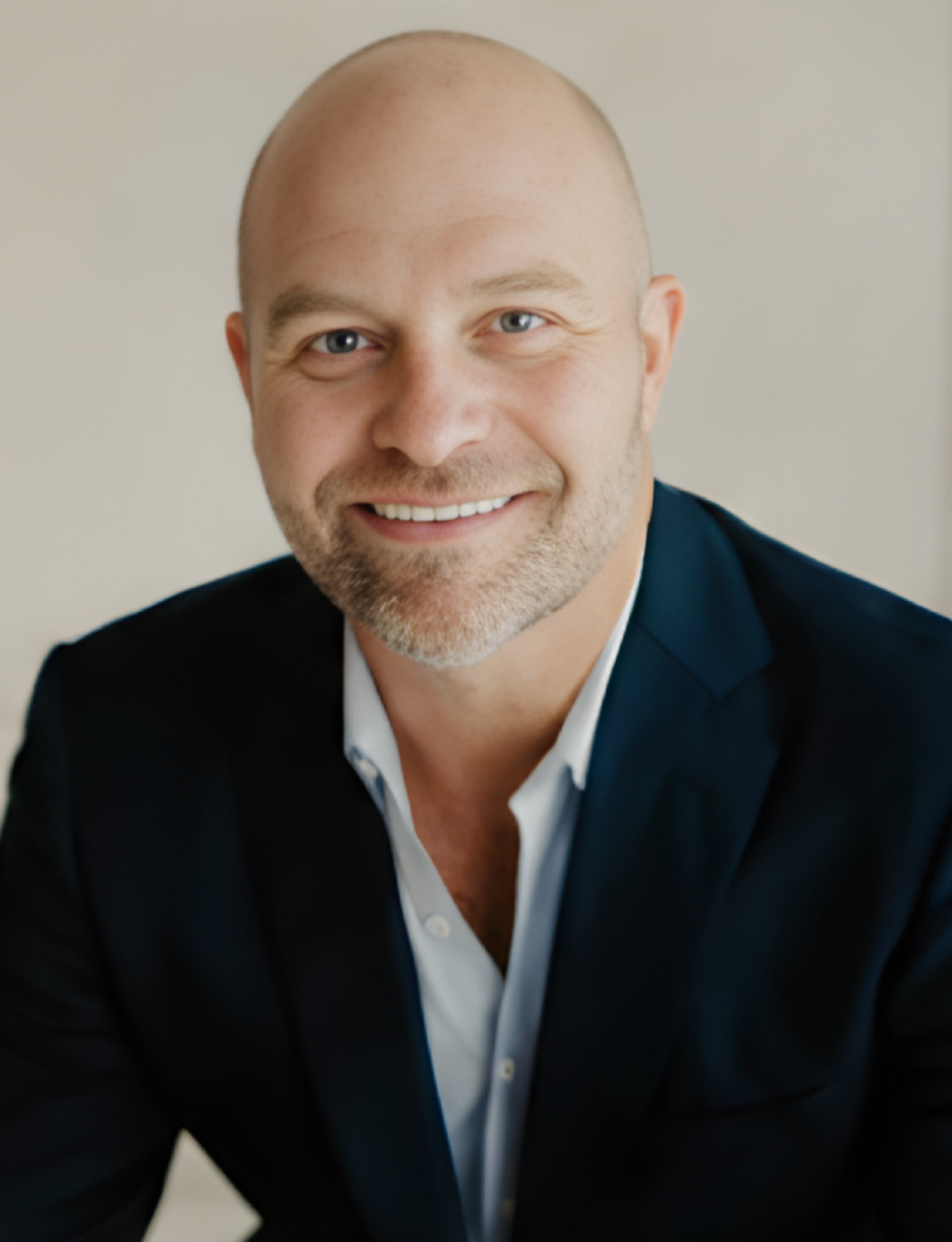 Man with a shaved head smiles, wearing a navy blazer over a light blue shirt.