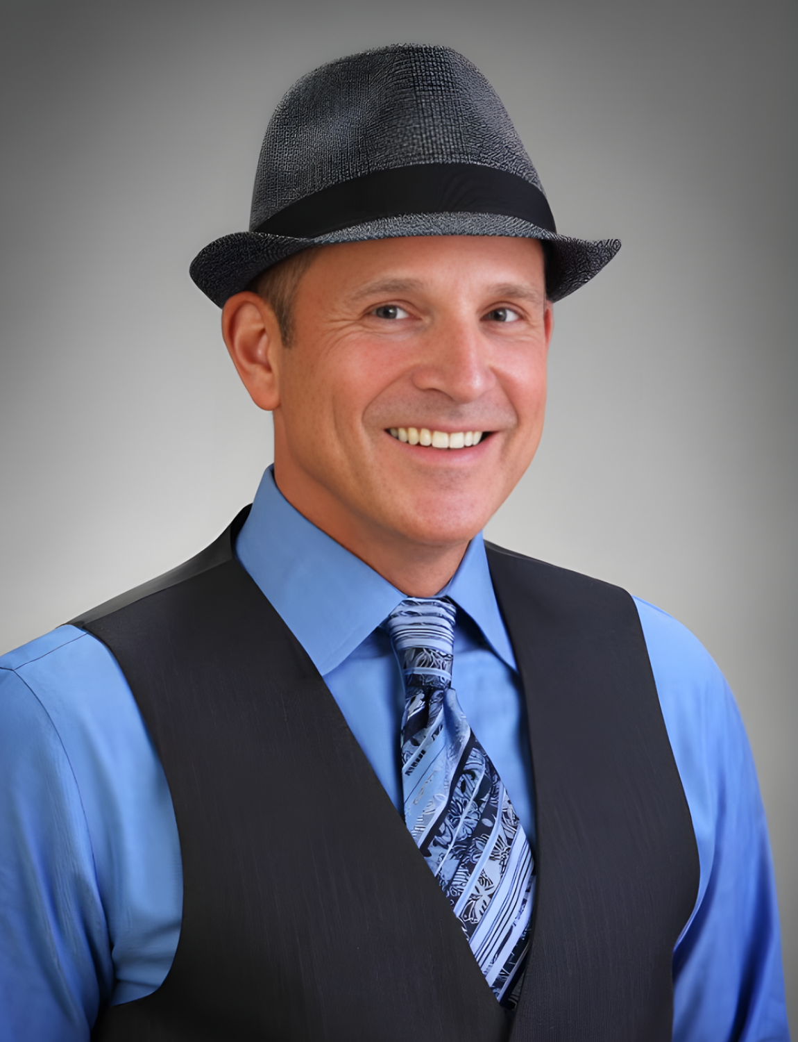 Man in blue shirt, vest, and fedora smiles at the camera.