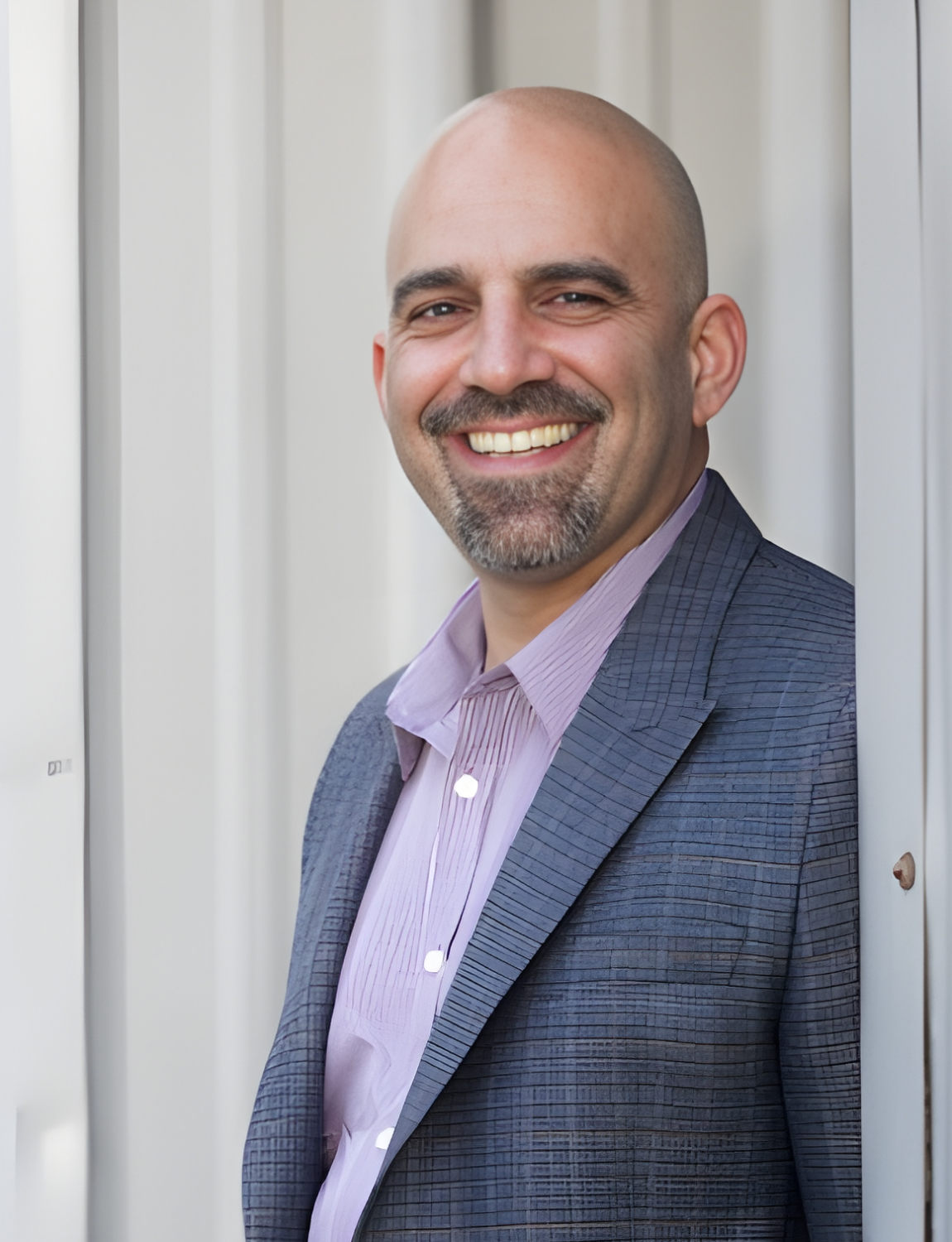 Bald man with a beard smiles, leaning against a white structure, wearing a blue blazer and pink striped shirt.