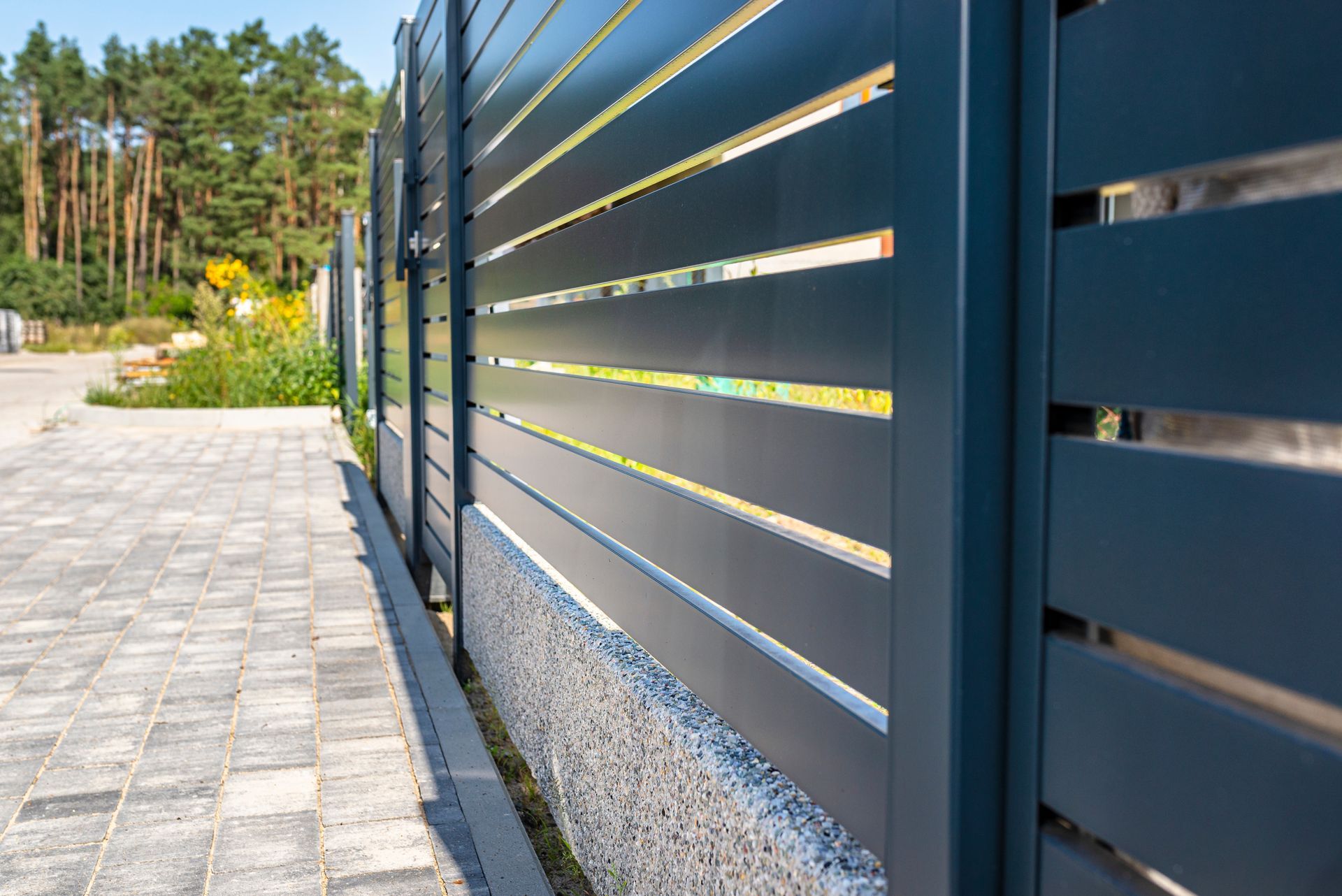 Close-up of a dark gray horizontal slat fence next to a brick walkway, with trees in the background in western NY