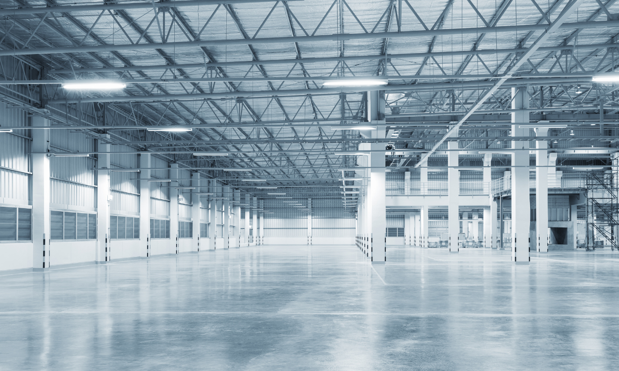 Empty, industrial warehouse interior with concrete floor, metal beams, and bright overhead lights.