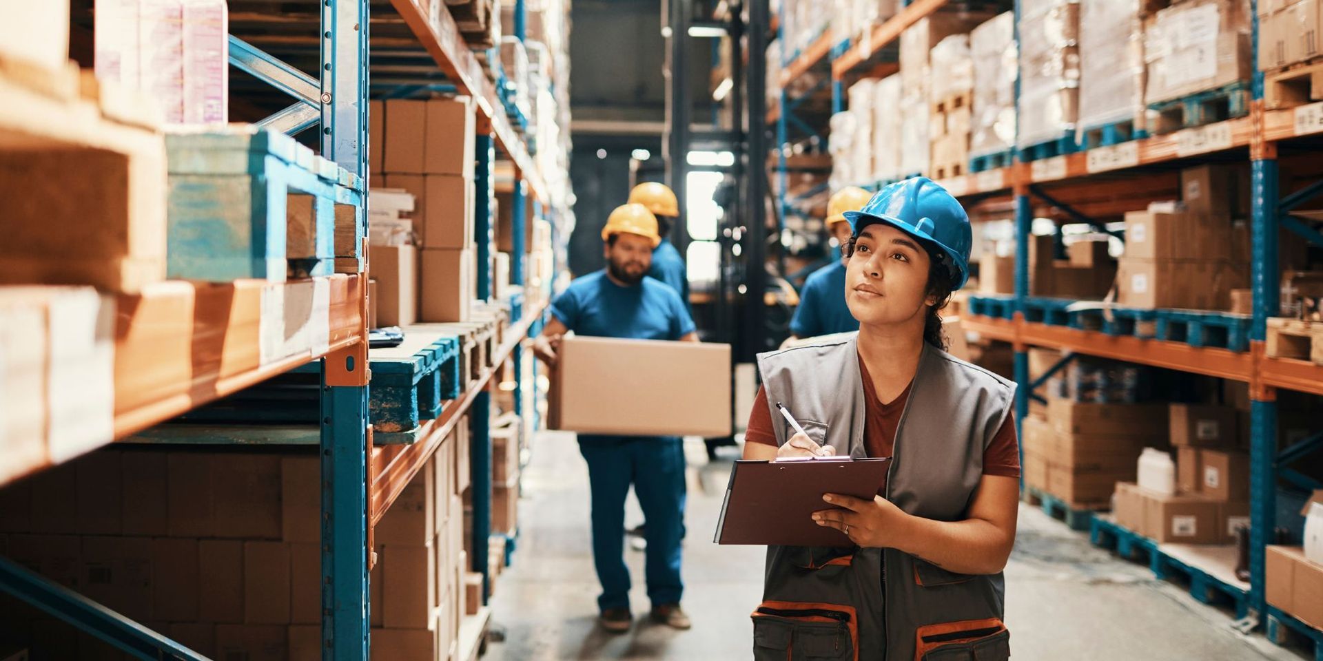 a woman is standing in a warehouse holding a clipboard and a box .