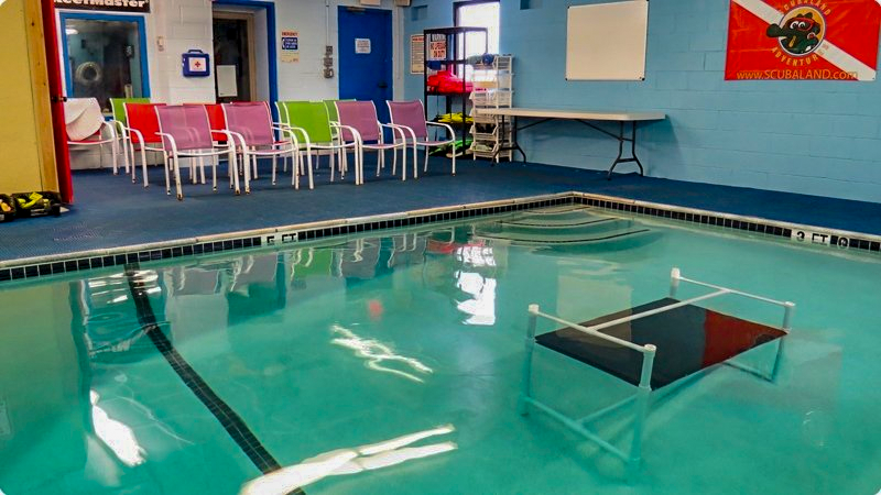 Indoor swimming pool with chairs, a table, and a diving platform.