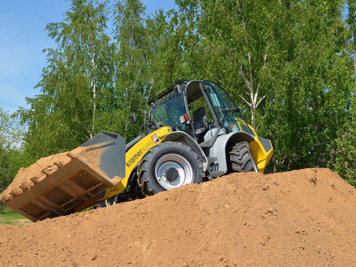 Yellow front loader dumping dirt on a sandy mound with green trees in the background