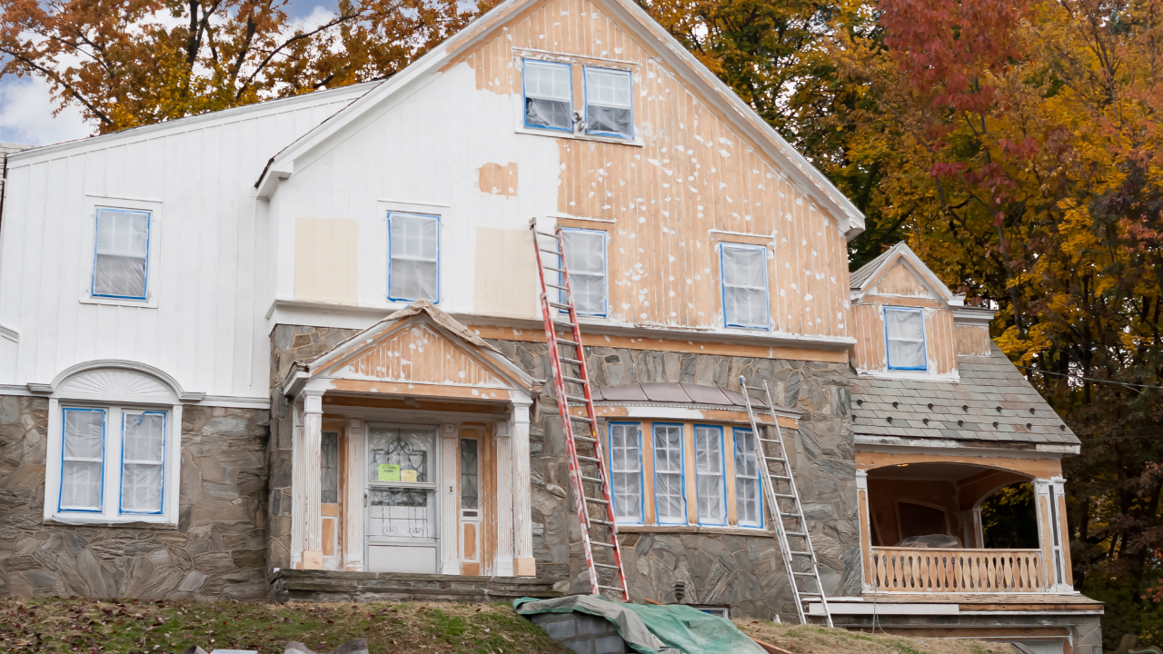 House being painted, half white and half weathered. Ladders and covered windows visible. Autumn foliage in background.