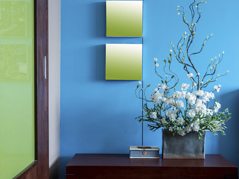 Blue wall with two square mirrors, floral arrangement, and small box on a wooden table.