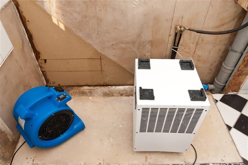 Blue fan and white dehumidifier drying a damp area with exposed walls.