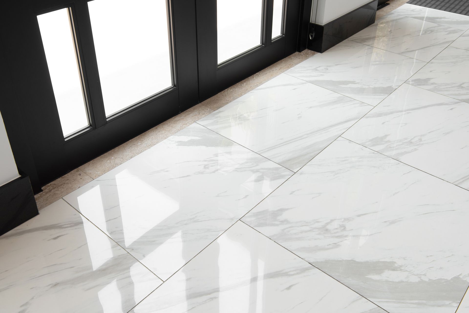 White marble tile flooring leading to a black door with clear glass panels; bright, reflective light.