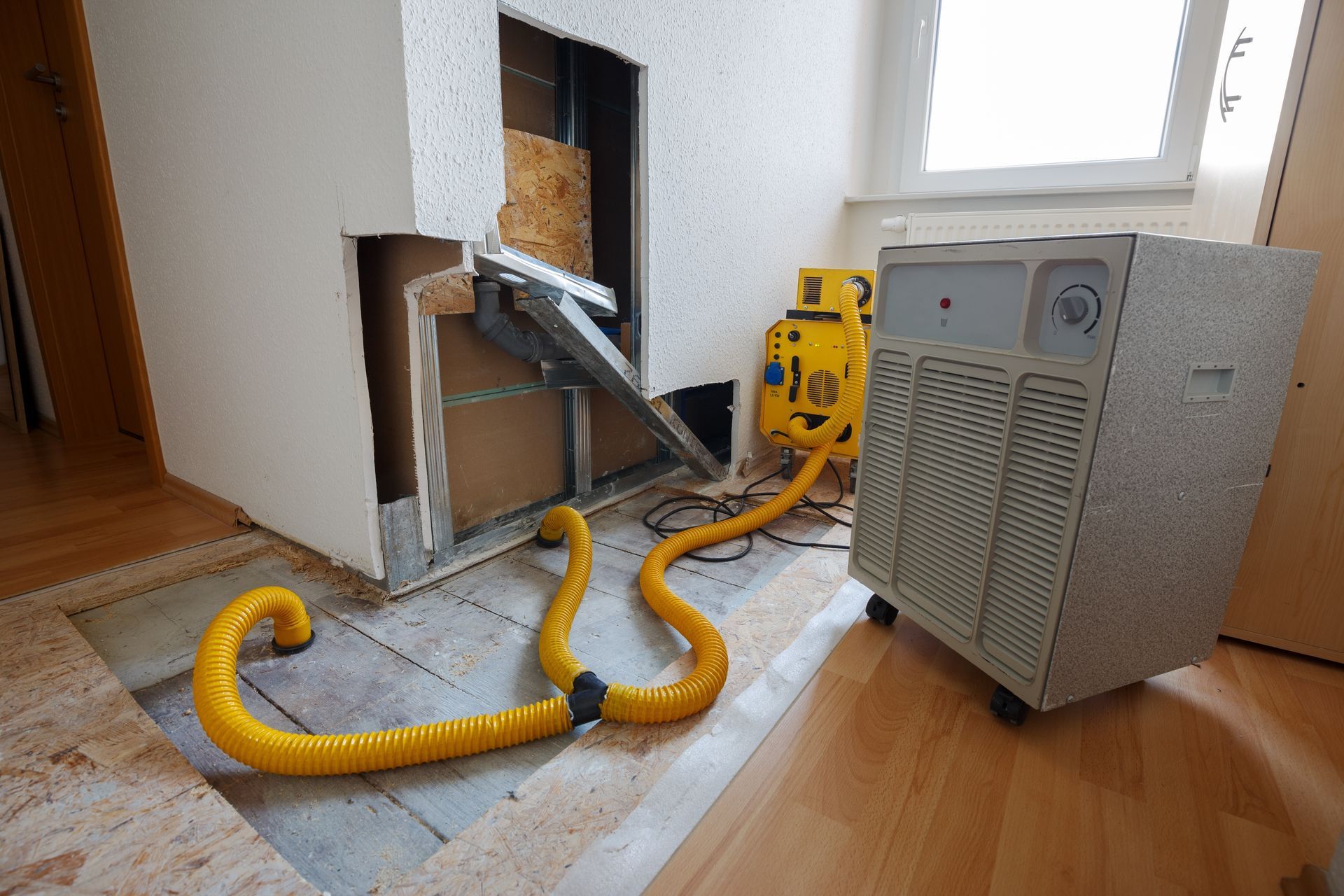 A yellow hose connects an air mover to a large dehumidifier. The equipment sits near a wall opening.