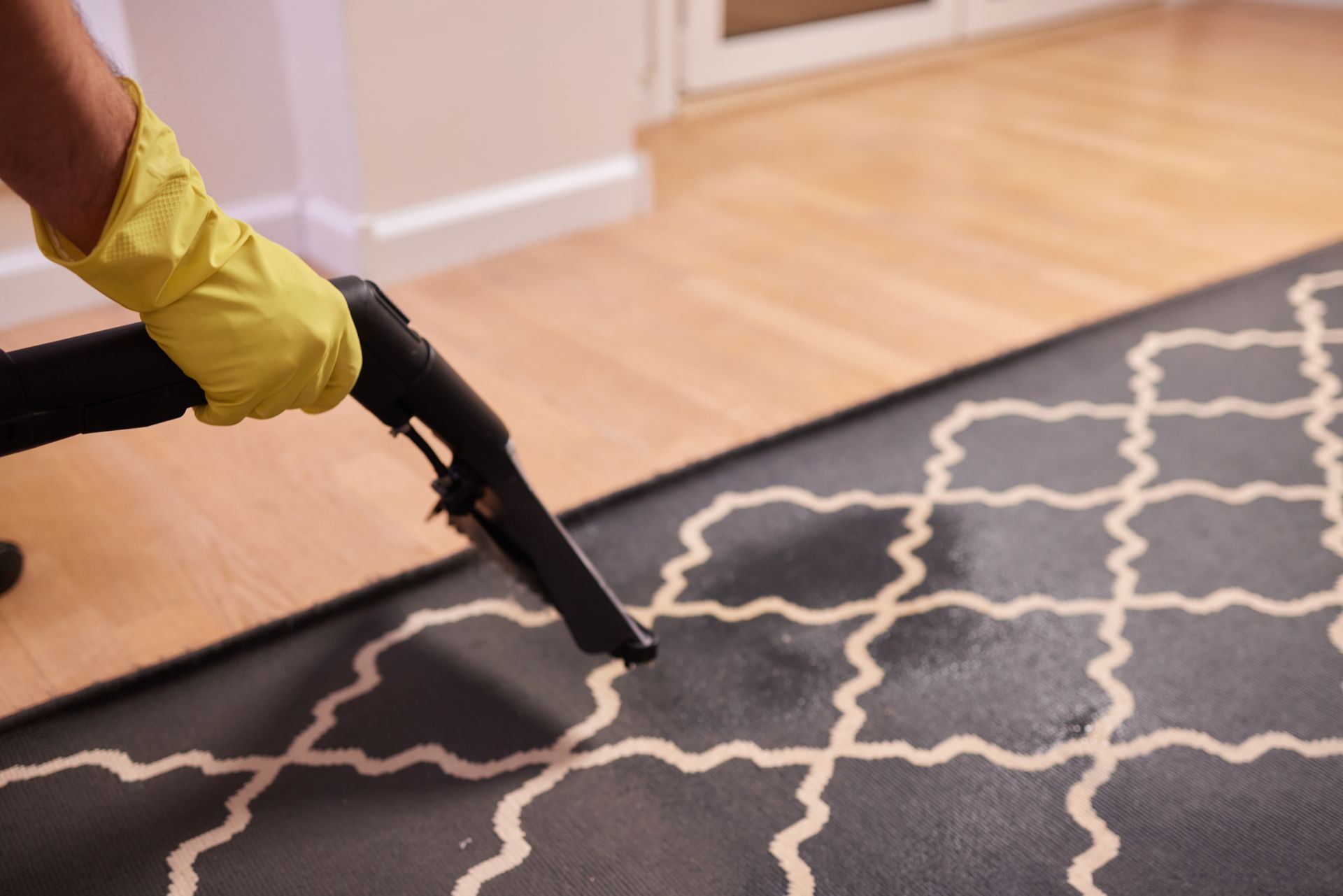 A gloved hand uses a cleaning tool on a patterned rug next to a wooden floor.