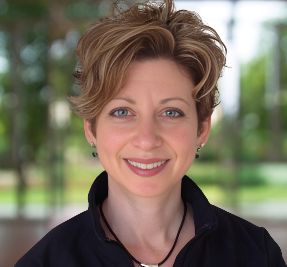 Woman with short, layered blonde and brown hair, smiling. Wearing black shirt with necklace, indoors.