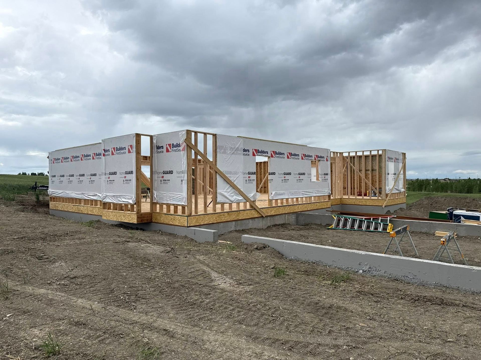 Framed house under construction on concrete foundation; wrapped in white; cloudy sky.