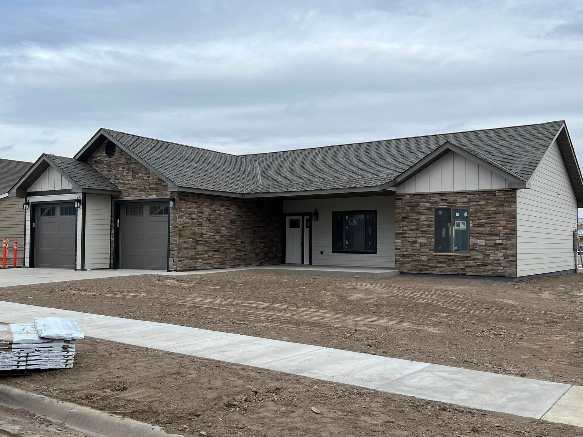 New home with stone and beige siding, two-car garage, and gray roof, on a cloudy day.
