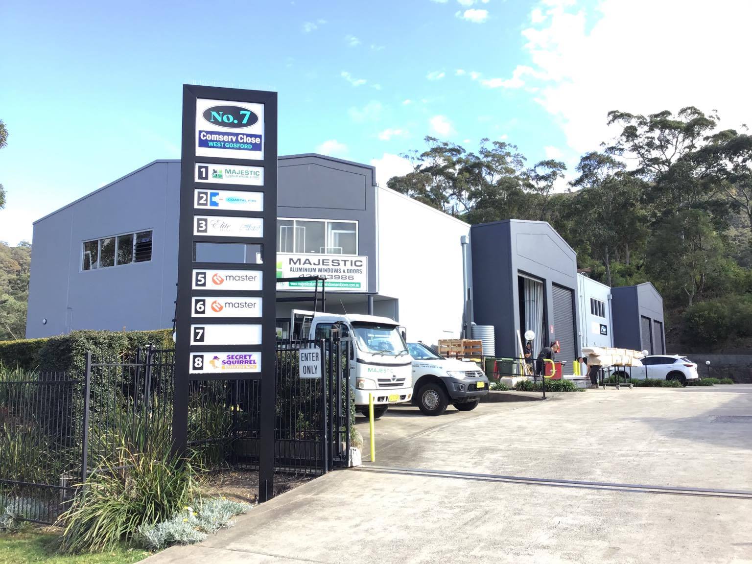 Building with multiple business signs, car, and trucks on a paved driveway. Trees in background.