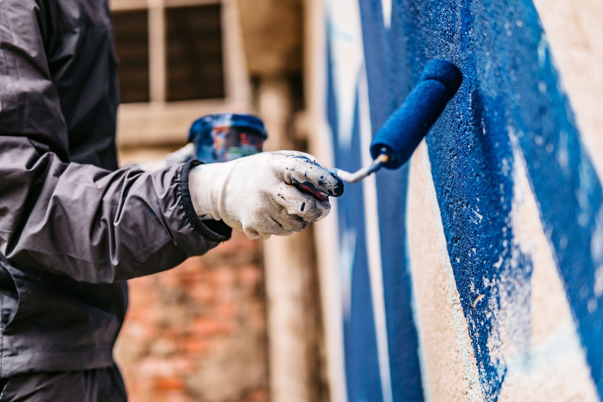 Close-up of a gloved hand as it holds a paint roller with blue paint as it paints a white wall.