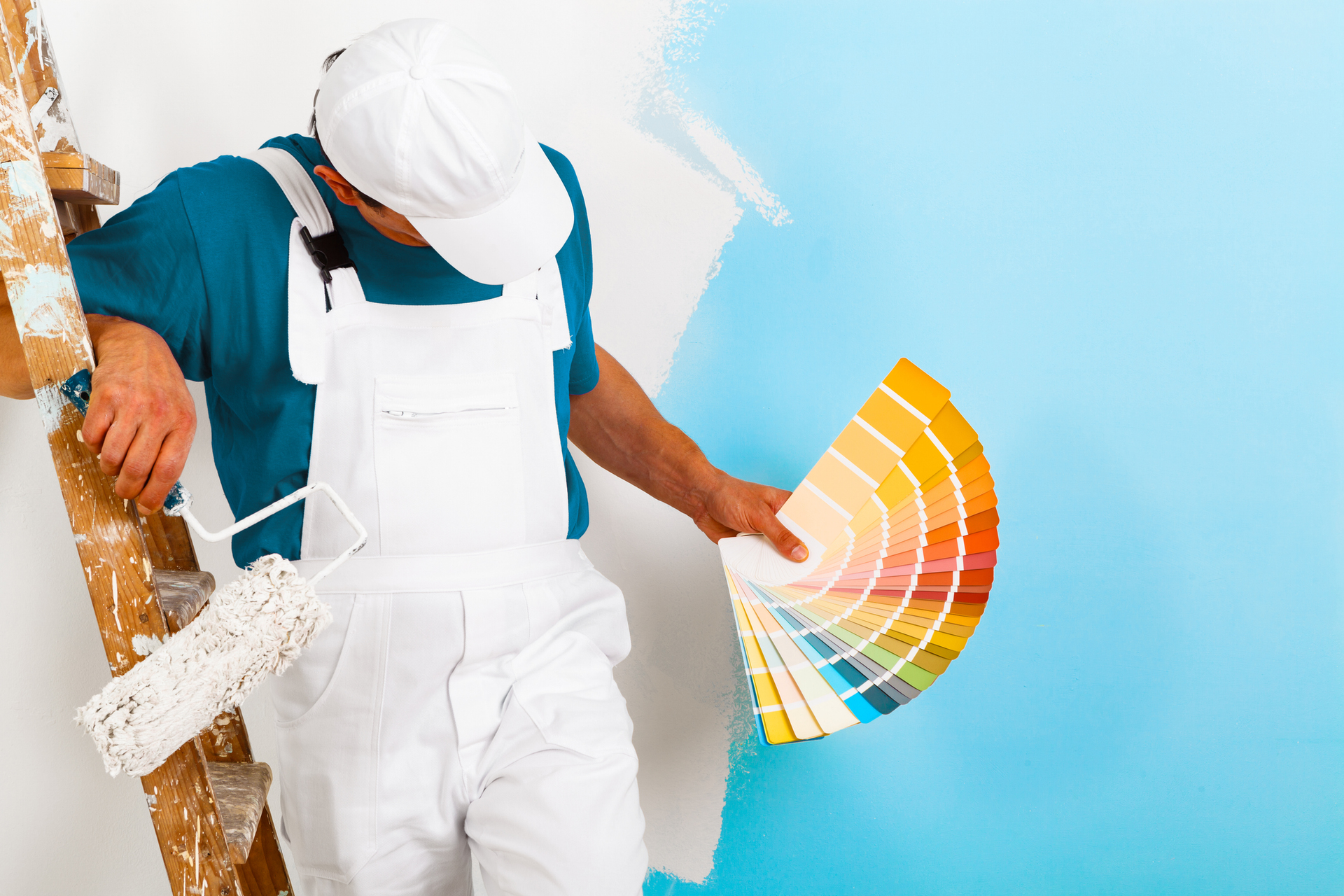 Painter with a paint roller and wooden ladder showing a colour palette, on a blue half-painted wall.
